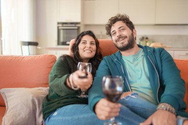 Genç bir çift oturma odasında bir anlık bağlantının tadını çıkarır. Adam, gri kapüşonlu adam ve kadın, koyu yeşil kazaklı, ikisi de kırmızı şarap bardakları tutuyor - romantik bir ilişki ve neşe paylaşımı.