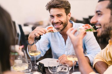 Bir grup genç rahat bir ortamda gurme pizzadan zevk alır. Şarap kadehleri yemek deneyimini güçlendirir. Arkadaşlar pizzanın tadını çıkarırlar.