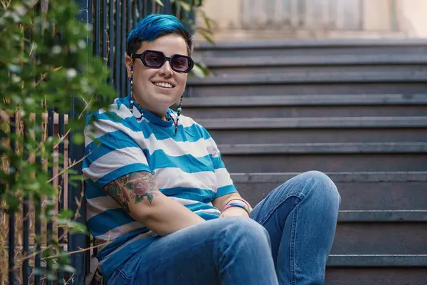 A genderfluid individual exuding relaxed confidence sits outdoors. Their blue hair, striped attire, and noticeable tattoo hint at their vibrant personality, while the urban backdrop provides a hint of city life.