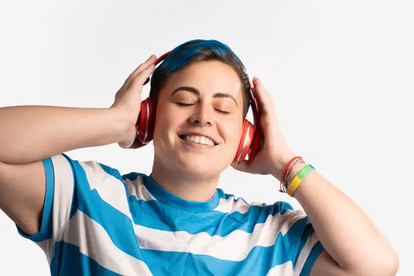 Portrait of a young, androgynous person with vibrant blue dyed hair, wearing headphones and a striped shirt. They are enjoying the music, showcasing a modern, casual style with nose piercing and colorful bracelets.