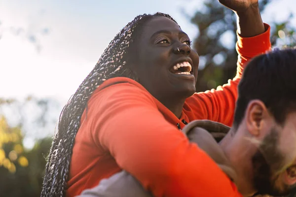 Joyful African descent woman with elongated braids gives a gleeful laugh while getting a piggyback ride from her partner outdoors, capturing a genuine moment of happiness