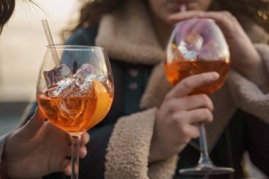 Portakal dilimleri ve buz küpleriyle süslenmiş spritz kokteylleri tutan ellerin yakın çekimi, rahatlama ve keyfi simgeliyor..