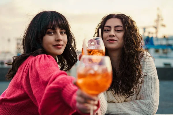 Two women share a serene moment by the harbor, toasting with spritz cocktails. The setting sun casts a golden hue, accentuating the bond they share against the maritime backdrop.
