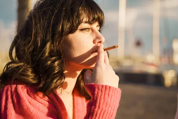 Young woman smoking by the sea - A young woman smokes by the sea, reflecting on youth\'s vices. The calm setting contrasts with her intense gaze, highlighting the complexities of modern indulgences.
