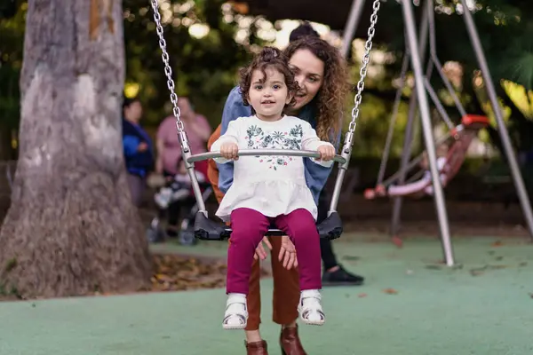 スイングで時間を楽しむ少女の心温まるイメージは 彼女の後ろに母親の存在と共に 彼らの笑顔と遊び心のある雰囲気は 家族が公園で共有する瞬間を強調しています — ストック写真