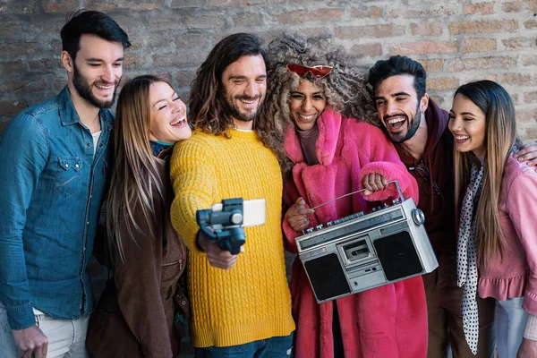 A group of young adults laugh against a brick wall, holding a retro boombox and filming a selfie video using a mobile gimbal. Their diverse appearances and joy blend modern youth with vintage vibes.