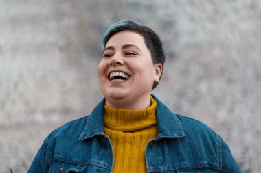 Saçında bir tutam deniz mavisi olan ikili olmayan bir kadın benzersiz kimliğini ve tarzını kucaklayarak mutluluk saçıyor. Parlak sarı balıkçı yaka kazağı ve sıradan kot ceketi moda anlayışına işaret ediyor..