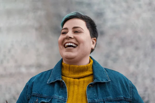 A non-binary woman with a dash of teal in her hair radiates happiness embracing her unique identity and style. Her vibrant yellow turtleneck and casual denim jacket hint at her trendy fashion sense.