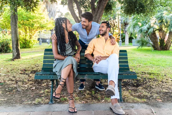 Three cheerful young adults, a diverse group, laughing and enjoying a sunny day on a park bench.