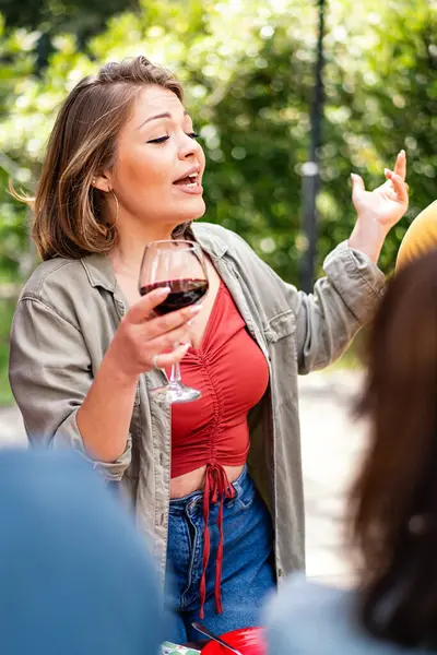 A woman animatedly tells a story during a social gathering, holding a glass of wine.