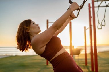 Odaklanmış, kızıl saçlı genç kadın güneş batarken açık hava spor salonunda süspansiyon antrenmanı kayışları kullanıyor..