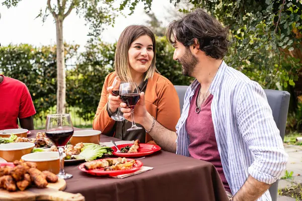 A couple engages in a friendly conversation over wine at an outdoor dining event, with a table full of various dishes in a lush garden setting.