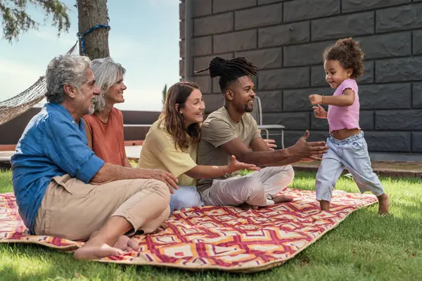 A multiracial family enjoys a picnic day as a toddler joyfully runs towards her parents and grandparents, capturing a moment of family bonding and happiness.
