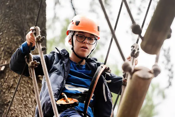 一个全神贯注的青少年戴着安全帽 带着安全带 参加了树木间富有挑战性的高绳课程 展示了敏捷和安全装备 — 图库照片
