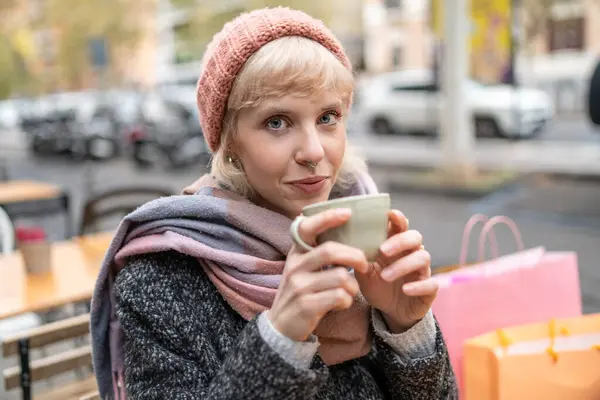 stock image A young blonde woman enjoys a warm cup of coffee on a city street, embodying a relaxed winter day.