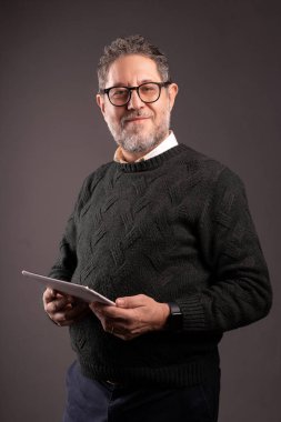 Gri bir arkaplanda duran, dijital tablet kullanan, günlük kıyafetli, olgun bir işadamı..