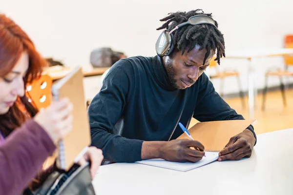 ヘッドフォンを持つ若いアフリカの男性学生は 意図的に勉強に吸収された教室の机でノートを書いています — ストック写真