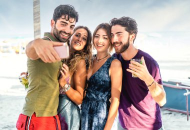 Neşeli bir grup arkadaş güneşli bir sahilde içki içerek selfie çekiyorlar..