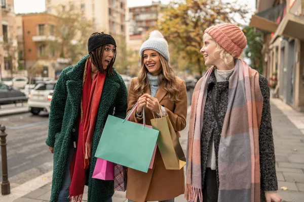Three young women stroll through city streets, smiling and chatting happily with numerous colorful shopping bags in hand, immersed in the vibrant autumn atmosphere.