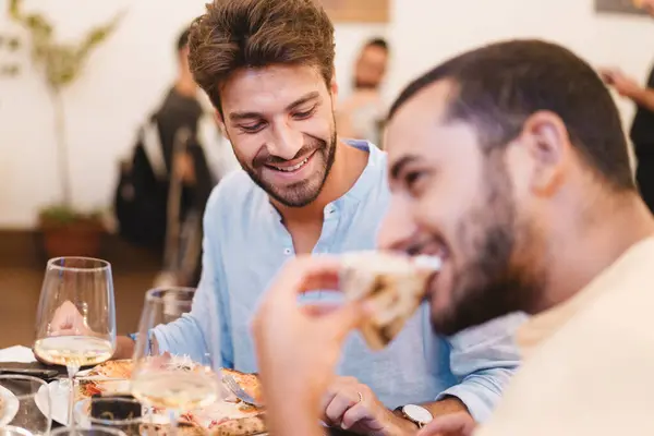 Two men laughing and sharing a meal with wine - Moments of friendship and joy at a casual dinner party.