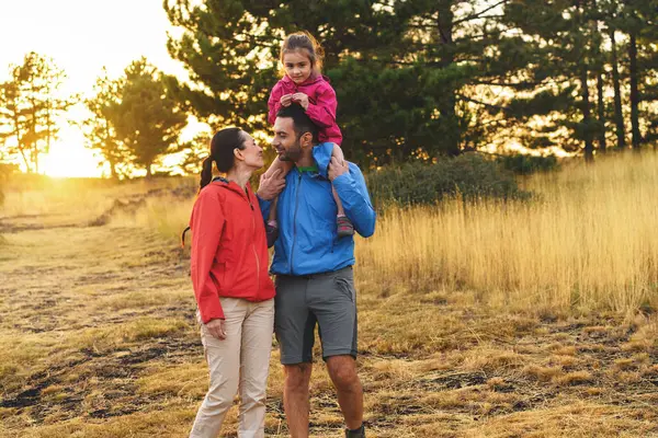 Family enjoying a hike with daughter riding on father\'s shoulders, sharing a moment in the warm sunset light.