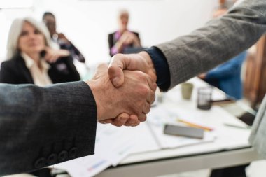 İş adamları el sıkışıyor - kapanış anlaşması, arka planda takım çalışması, başarılı pazarlıklar - arka planda diğer iş adamlarıyla modern ofis ortamları