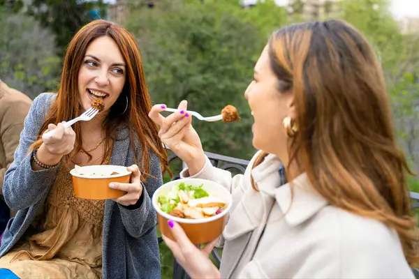 İki kadın, geri dönüşümlü kağıt kaseler ve gübreli çatallar kullanarak çevre dostu çatallarla öğle yemeği yiyor. Açık havada, sürdürülebilir yiyecekler ve mutlu anlar..