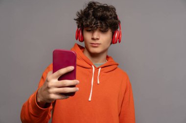 Turuncu kapüşonlu ve kırmızı kulaklıklı bir genç, akıllı telefonlu bir selfie çekiyor, gündelik moda, modern teknoloji, izole edilmiş bir geçmiş, gençlik kültürü