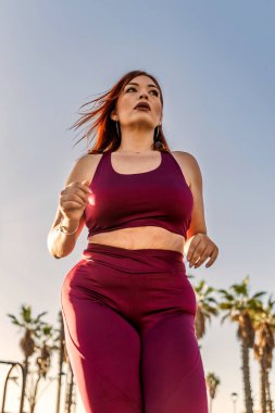 Kestane rengi spor kıyafetleriyle dışarı koşan iri yarı bir kadın azim ve zindelik motivasyonu, sağlıklı yaşam tarzı, parlak ve güneşli bir gün, aktif ve dinamik bir hareket sergiliyor.