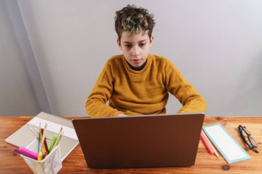 Hardal süveter giyen genç bir çocuk evde dizüstü bilgisayarda çalışırken dikkati dağılmış görünüyor. Okul malzemeleri de masanın üzerinde. Bu ifade, ev ortamında derin düşüncelere dalmayı ya da hayal kurmayı öneriyor..