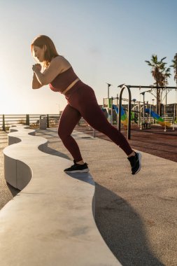 Genç sporcu kadın, deniz kenarındaki bir parkta beton bir bankta bacaklarını çalıştırıyor.