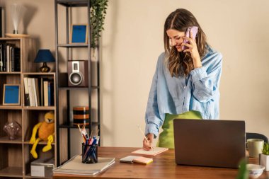 Ofiste not alırken telefonla konuşan profesyonel bir kadın. Çoklu görev ve odaklanma - uzak çalışma, ev ofis kurulumu, verimlilik sahnesi, modern çalışma alanı