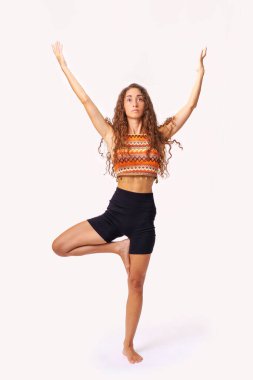 Yogada ağaç pozu veren genç bir kadın. Renkli bir etek giyiyor. Beyaz arka planda modern fitness uygulamasında denge, güç ve kültürel çeşitlilik gösteriliyor.