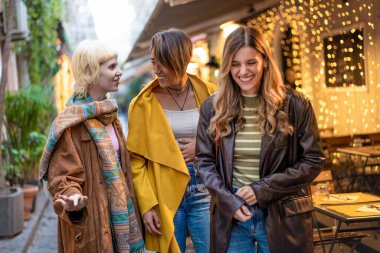 Farklı etnik kökenlere sahip üç zarif genç kadın açık hava kafesinde keyifli bir anı paylaşıyor. Görüntü, arkadaşlığı, çeşitliliği ve çağdaş modayı sıcak ışıklandırmayla büyüleyici bir şehir ortamında yakalar..