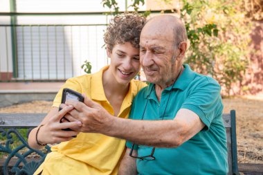 Genç kıvırcık saçlı torun dışarıda bankta otururken yaşlı büyükbabanın akıllı telefon kullanmasına yardım ediyor. Nesiller arası öğrenme ve teknoloji benimseme kavramı.