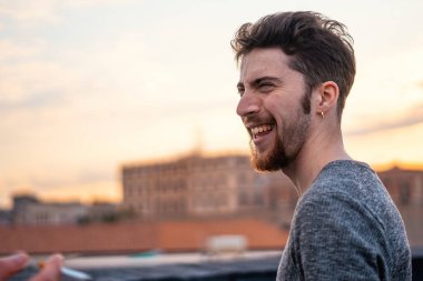 Gün batımında şehrin çatısında gülen neşeli, sakallı bir adamın samimi portresi. Sıcak akşam ışığında çekilen doğal, hakiki duygu şehir yaşam tarzını ve mutluluğu gözler önüne seriyor..