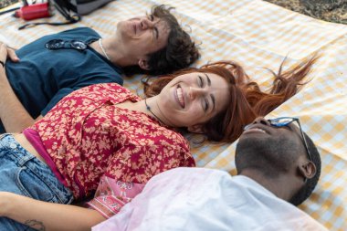 Bir grup çok kültürlü genç arkadaş dışarıda kareli bir battaniyenin üzerinde yatıyorlar. Gülümsüyorlar ve birlikte kaygısız bir anın tadını çıkarıyorlar, doğal bir ortamda arkadaşlık ve çeşitlilik gösteriyorlar..