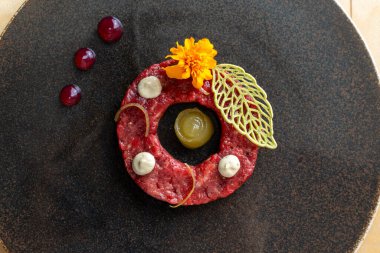 Top view of elegant beef tartare ring with egg yolk, marigold flower, and decorative green leaf. Modern fine dining presentation on dark plate with red sauce dots. Luxury restaurant dish. clipart