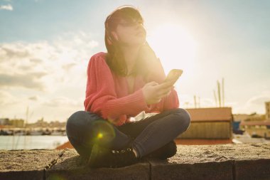 Altın saatinde mobil teknoloji kullanan genç bir kadın. Z jenerasyonu dijital doğal ışık efektiyle dışarıda rahatlama, otantik yaşam tarzı anı