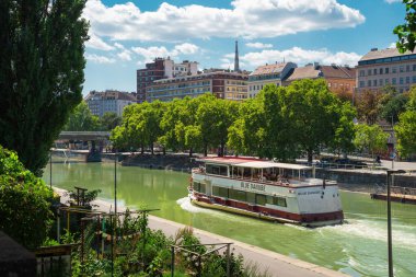 VIENNA, AUSTRIA - 11 Ağustos 2024: Donaukanal boyunca arka planda St. Stephen 's Katedrali kulesi olan turist botu Blue Tuna seyahat ediyor. Yaz şehir manzarası yeşil nehir kıyısı ve şehir binalarını gösteriyor (182 caratteri)