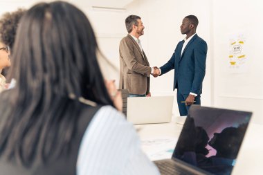 Çeşitli iş adamları modern ofis ortamında gözlem yapan takım üyeleriyle şirket toplantısı sırasında el sıkışıyor.