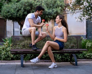 Genç çift, şehirde yaz öğleden sonra bankta otururken birlikte dondurma yiyorlar.
