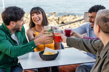 Sahil kenarındaki barda içkiyle kutlama yapan çok kültürlü genç arkadaşlarla samimi bir an. Yaz aylarında yapılan sosyal toplantılarda doğal neşe ve kahkaha ifadeleri.