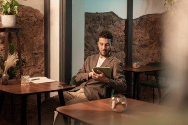 Çağdaş restoranda taştan duvarları olan dijital tablet kullanan bej ceketli genç bir profesyonel. Modern iş adamı, doğal dekorlu zarif bir kafede çalışıyor..