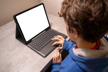 Masasında klavyesi olan tablet kullanan genç bir çocuk. Cihaz ekranı boş ve beyazdır, çevrimiçi öğrenme, dijital eğitim veya günlük tarama önerisi.