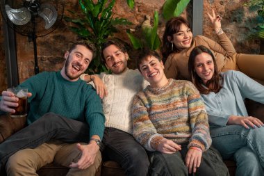 Neşeli bir grup arkadaş bir kanepede oturuyor, gülümsüyor ve rahat bir kafede, yeşil ve sıcak dekorlarla çevrili, arkadaşça ve neşeli bir anın tadını çıkarıyorlar..