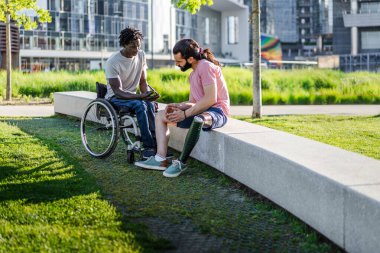 İki arkadaş, biri protez bacaklı ve diğeri tekerlekli sandalye kullanıyor, modern bir şehir parkında beton bir bankta oturuyor. Anlamlı bir sohbete dahil olmak, bağlantı kurmak, güçlendirmek.