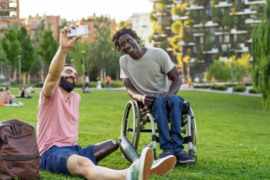 İki arkadaş, biri protez bacaklı, diğeri tekerlekli sandalye kullanıyor yeşil bir şehir parkında selfie çekerken gülümsüyor. Görüntü, modern bir açık hava ortamında kapsayıcılığı, dostluğu ve olumlu bağlantıları vurguluyor..