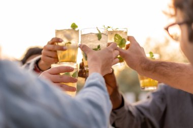 Yakın plan bir arkadaş bardaklarını nane yapraklarıyla süslenmiş ferahlatıcı kokteyllerle tokuşturuyor, gün batımını dışarıda kutluyorlar. Arkadaşlık, kutlama ve yaşam tarzı temaları için mükemmel..