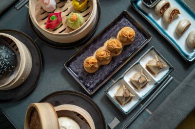 Bambu buharlı buharlı ve seramik tabaklarda servis edilen çeşitli Dim Sum manzarası. Renkli hamur köfteleri, mantarlı paketler ve modern karanlık masa ortamındaki diğer lezzetleri içerir..